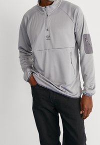 Muž v světle šedém pulovru Adidas s čtvrtinovým zipem a kapsou na zip na rukávu, v tmavých kalhotách, stojí s jednou rukou v kapse.