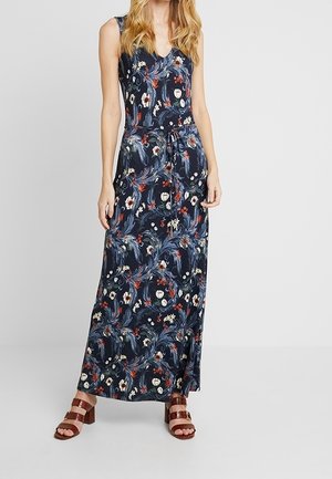 Mujer con un vestido largo sin mangas de color azul marino con estampados florales y un lazo en la cintura, combinado con sandalias marrones de tacón ancho.