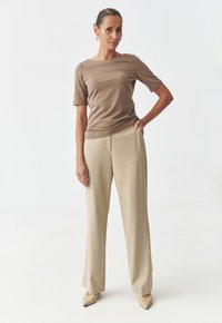 Haut marron à manches courtes avec un décolleté droit, associé à un pantalon large beige clair. Modèle portant des chaussures à bout pointu nude.