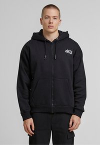Sweat à capuche noir avec fermeture éclair, fabriqué en tissu épais, doté d'une poche avant, d'une capuche avec cordon de serrage et d'un petit logo blanc sur la poitrine.