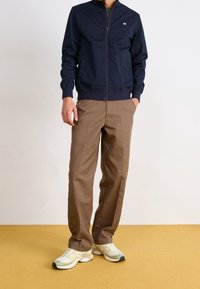 Granatowa kurtka na zamek z kołnierzem stojącym, brązowe spodnie o luźnym kroju oraz jasne buty sportowe. Prosty design bez wyraźnych wzorów czy zdobień.