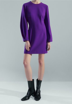 Femme portant une mini robe à manches longues violette associée à des bottes à talons noires mi-mollet, debout devant un fond uni.