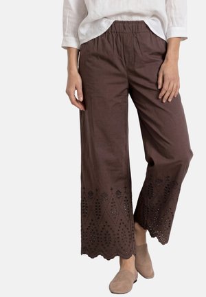 Kvinde iført brune bukser med vide ben og broderede bølgede kanter, kombineret med beige slip-on sko og en hvid top.