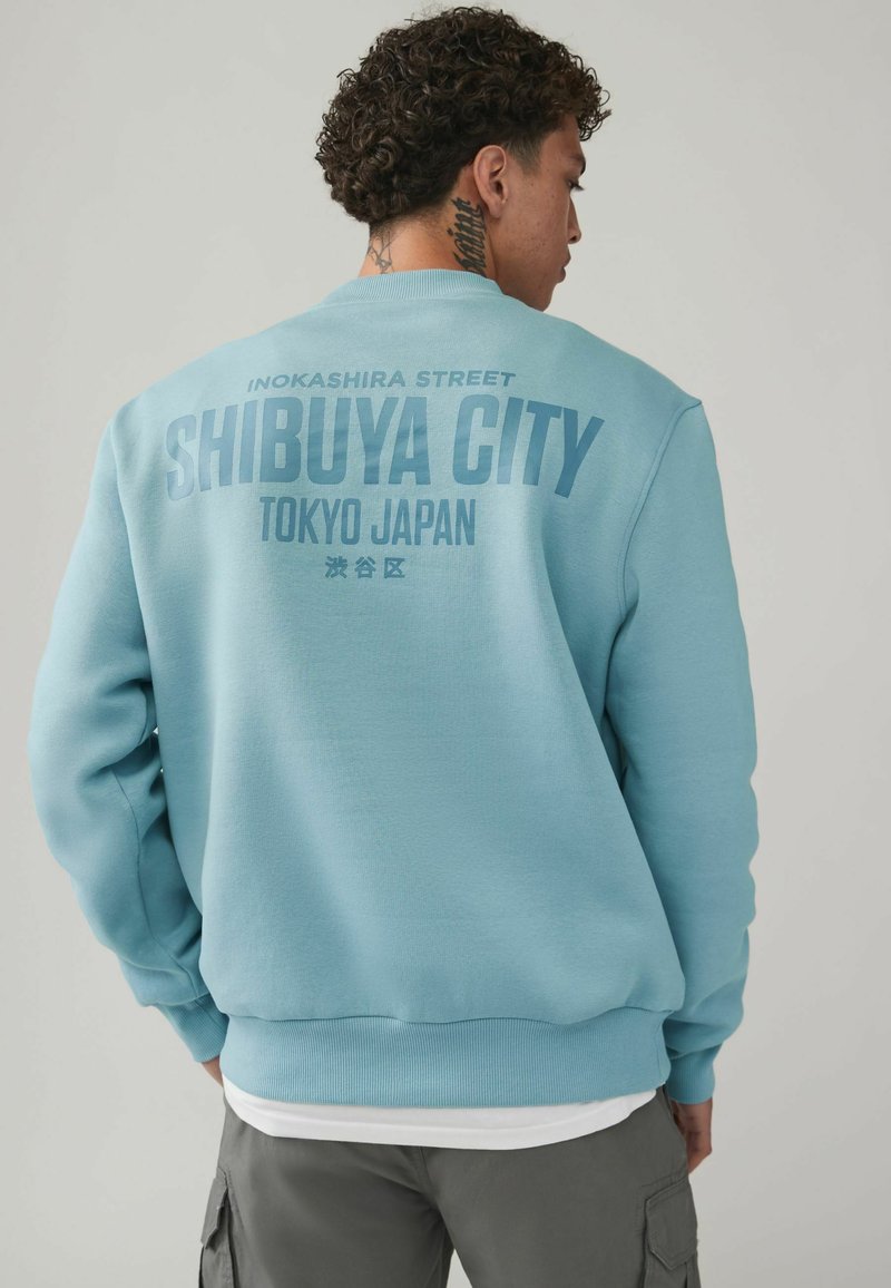 Tyrkysová mikina s rebrovanými manžetami a spodným lemom, na chrbte s veľkým textom: "SHIBUYA CITY TOKYO JAPAN" v kontrastnej modrej.
