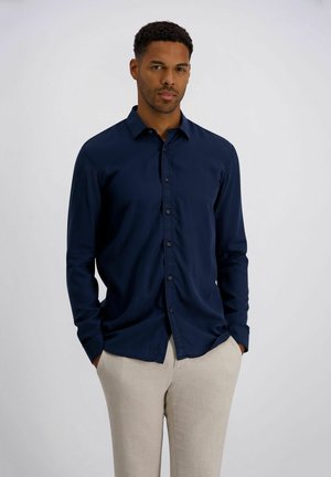 Mann mit Händen in den Taschen, trägt ein marineblaues Hemd mit Knöpfen und beige Hose vor einem schlichten hellen Hintergrund.