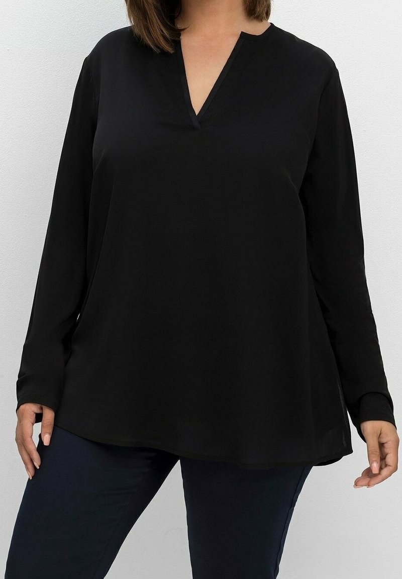 Femme portant un chemisier noir ample à manches longues avec col en V et un pantalon sombre, debout devant un fond clair uni.