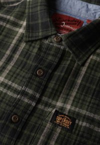 Groen en zwart geruite flanellen shirt met een kraag, bruine knopen en een zwart en oranje "Superdry" label op de borst. Gelaagd stof.
