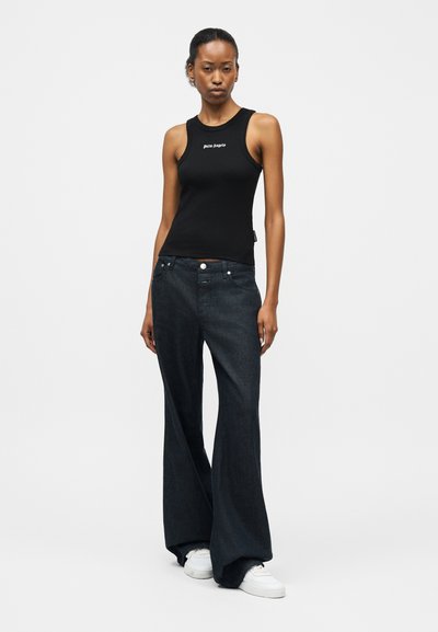 Γυναίκα που στέκεται φορώντας μαύρο αμάνικο τοπ με το λογότυπο "Palm Angels", φαρδιά μαύρα τζιν παντελόνια και λευκά αθλητικά παπούτσια σε λευκό φόντο.