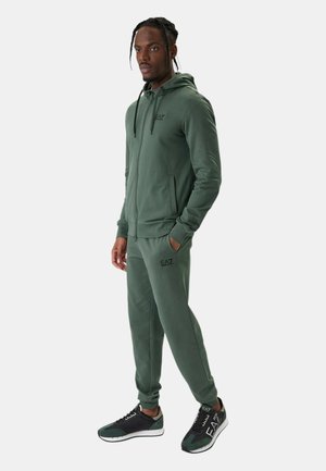 Uomo in piedi che indossa giacca verde con cappuccio e zip e pantaloni coordinati con scarpe da ginnastica nere e verdi, mani parzialmente nelle tasche, guarda avanti.