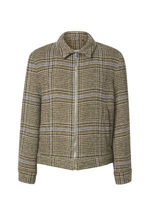 Jeune homme debout portant une veste zippée à carreaux beige, un jean bleu foncé et des bottes en cuir noir sur un fond clair uni.