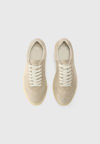 Beige suede sneakers with white laces, round toe, and a textured sole. Branding visible on the tongue and side. Smooth interior lining.