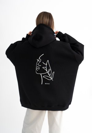 Zwarte hoodie met een witte lijntekening van een gezicht en bladeren op de achterkant, gemaakt van zacht materiaal en met een relaxte pasvorm.