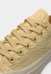 Baskets jaune clair avec un matériau texturé et froissé, lacets plats et œillets circulaires ; elles possèdent une semelle en caoutchouc blanche.