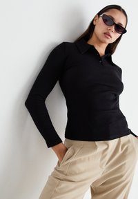 Camisa polo negra de manga larga con textura acanalada y un cuello, combinada con pantalones plisados en un tono beige claro. Modelo porta gafas de sol oscuras.