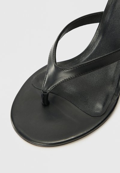 Tongs en cuir noir présentant un design à l'arrière du pied, avec une semelle plate et une texture lisse. Les lanières sont minimalistes et fixées de manière sécurisée.