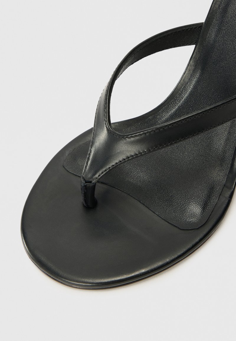 Chancla de piel negra con un diseño de tira para los dedos, suela plana y textura suave. Las correas son minimalistas y están fijadas de manera segura.
