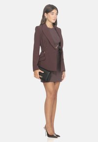 Burgunderfarbener maßgeschneiderter Blazer mit goldenen Accessoires und Seitentaschen, getragen über einem engen braunen Kleid. Das Model hält eine schwarze Clutch und trägt spitze Absätze.