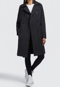 Trench-coat noir à double boutonnage, grands boutons et col large, assorti à des leggings noirs et des baskets blanches. Moderne et élégante.