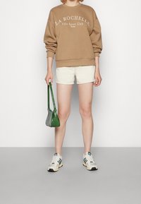 Osoba s hnědou mikinou "La Rochelle", bílými šortkami a bílými teniskami Adidas, držící malou zelenou kabelku na bílém pozadí.