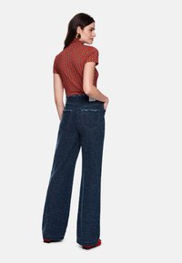 Rotes Oberteil mit Muster und kurzen Ärmeln, kombiniert mit hoch taillierter, weiter blauer Jeans aus strukturiertem Stoff mit Gesäßtaschen. Rote Schuhe.