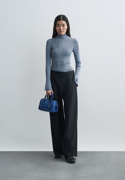 Woman wearing a long-sleeve striped top and wide black pants, holding a blue patterned handbag, standing against a plain gray background.