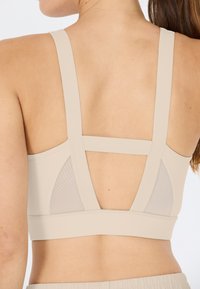 Beige urheilusetti, jossa on leveät hihnat, verkkomallinen takapaneeli ja saumatonta muotoilua. Sujuva tekstuuri, minimalistinen ulkonäkö ja mukava istuvuus.