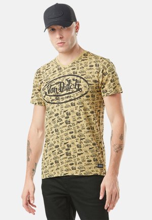 Homme portant une casquette noire et un t-shirt graphique jaune Von Dutch avec des textes et gribouillis noirs partout, associé à un pantalon noir.