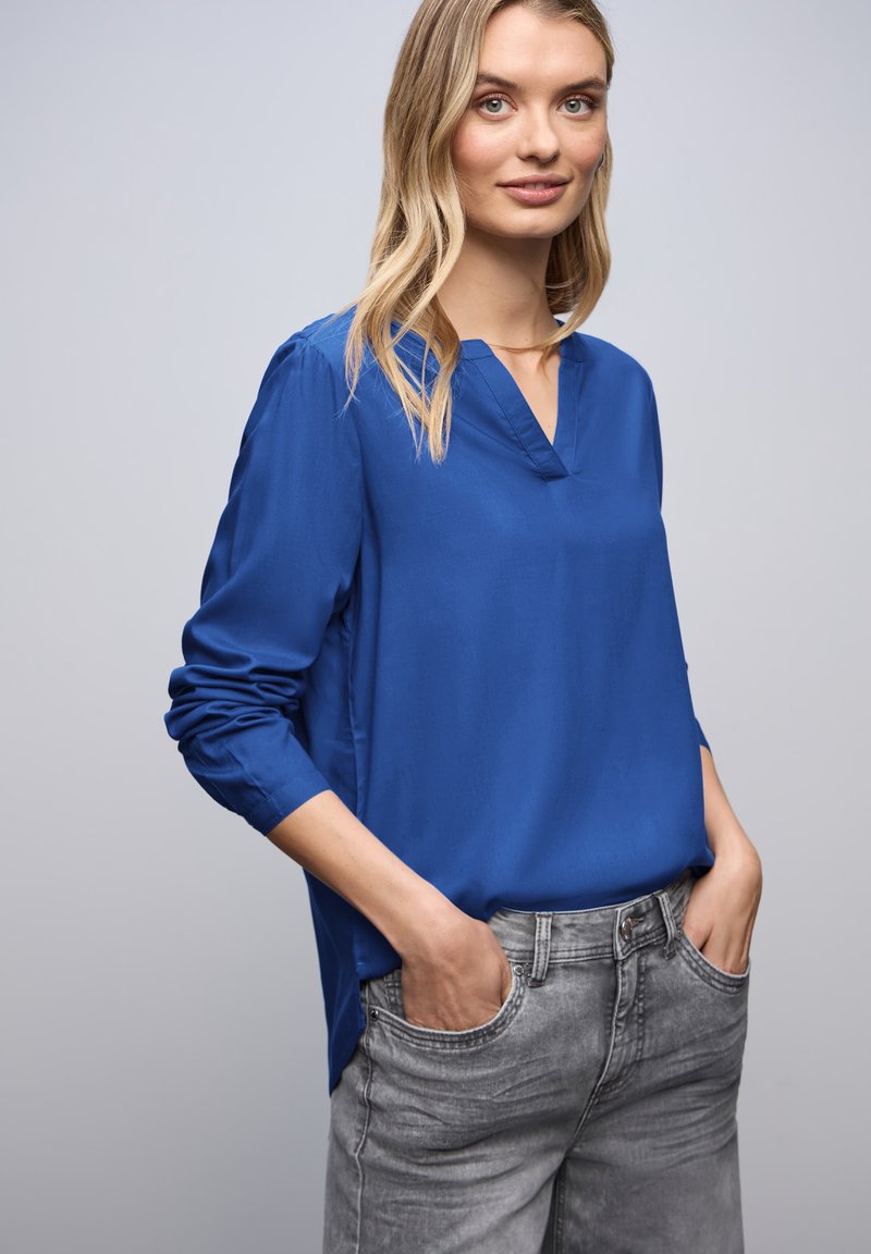 Blå langærmet bluse med V-hals, lavet af glat stof. Den har oprullede ærmer og er parret med grå jeans.