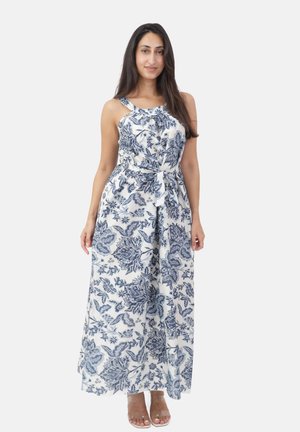 Mujer vestida con un vestido maxi blanco sin mangas con estampados florales azules, cintura ceñida con cinturón y sandalias color canela abiertas en los dedos, de pie contra un fondo liso.