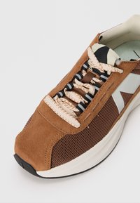 Braune Wildleder-Sneaker mit Netzdetails, cremefarbenen Schnürsenkeln, schwarzen und weißen Akzenten sowie einer schlanken, runden Form. Verfügt über eine strukturierte Sohle.