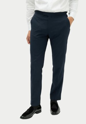Man som bär mörkblå formella byxor, svarta toffelloafers och en vit långärmad skjorta med händerna i fickorna.