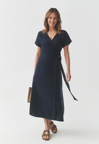 Abito avvolgente navy con maniche corte, annodato in vita e con gonna lunga. Abbinato a sandali marroni e una borsa piccola. Tessuto a trama liscia.