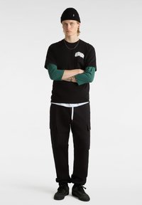 Camiseta negra de manga corta con un gráfico blanco, superpuesta sobre una camiseta de manga larga verde. Pantalones cargo negros y zapatillas negras completan el look.