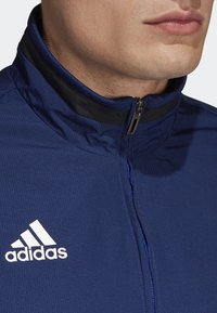 Mörkblå träningsjacka med dragkedja i kragen, med svarta detaljer, strukturerad tyg och en vit Adidas-logotyp på v änster sida.