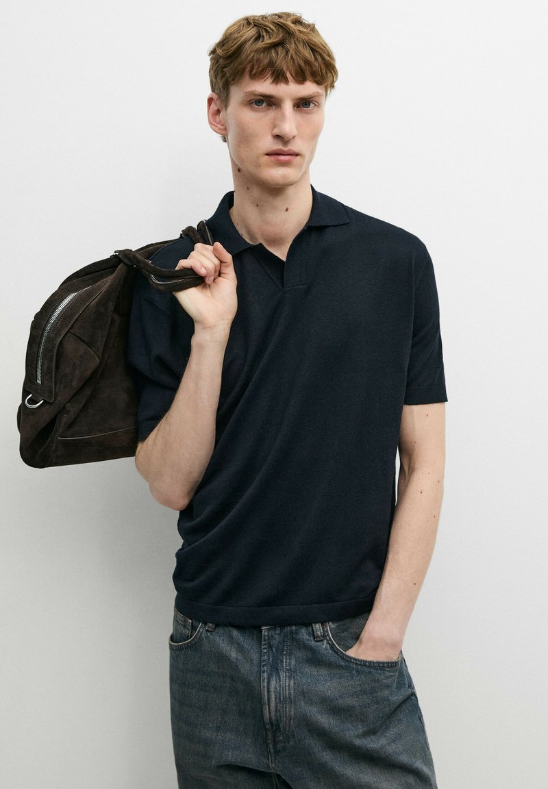 Giovane uomo con polo nero e jeans, che tiene uno zaino in pelle scamosciata marrone scuro sulla spalla, su sfondo bianco uniforme.