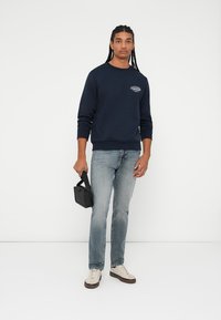 Mørkeblå crewneck sweatshirt i bomullsblanding, med en logo på brystet. Kombinert med lyseblå jeans og kremfargede sneakers, holder en svart veske.