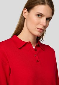 Rotes Polohemd mit Kragen, ausgestattet mit drei Knöpfen und einem strukturierten Stoff, präsentiert an einem Modell vor einem neutralen Hintergrund.