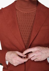 Rustfarvet blazer med en tekstureret, strikket turtleneck under. Blazeren har en klassisk revers og en forlommer, fastgjort i taljen.