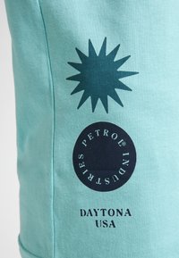 Camiseta de algodón turquesa con un estallido de estrellas azul oscuro y un logo circular que dice "PETROL INDUSTRIES DAYTONA USA". Textura suave.
