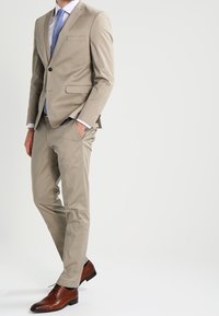 Man staat met zijn handen in zijn zakken, gekleed in een beige pak, lichtblauwe shirt, blauwe stropdas en bruine nette schoenen op een witte achtergrond.