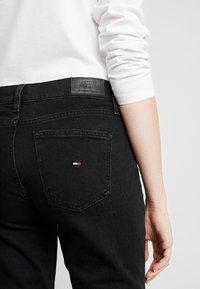Jeans negros con un detalle de bolsillo visible y una etiqueta de Tommy Jeans, usados con una camisa blanca de manga larga, enfocados en la vista posterior.