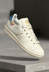 Valge nahast Adidas Stan Smith sneaker, millel on värviline mustriga kannaaktsent, kuldne bränding ja ventilatsiooniavad.