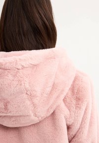 Bata de baño de felpa rosa con textura suave y peluda; presenta un corte holgado y una gran capucha con detalles de costura en la parte posterior.