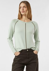 Kobieta o długich blond włosach, ubrana w jasnozielony sweter z ciemnymi pionowymi pasami i ciemnobrązowe spodnie, z rękami w kieszeniach, patrząca na bok.