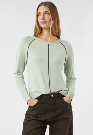 Kobieta o długich blond włosach, ubrana w jasnozielony sweter z ciemnymi pionowymi pasami i ciemnobrązowe spodnie, z rękami w kieszeniach, patrząca na bok.