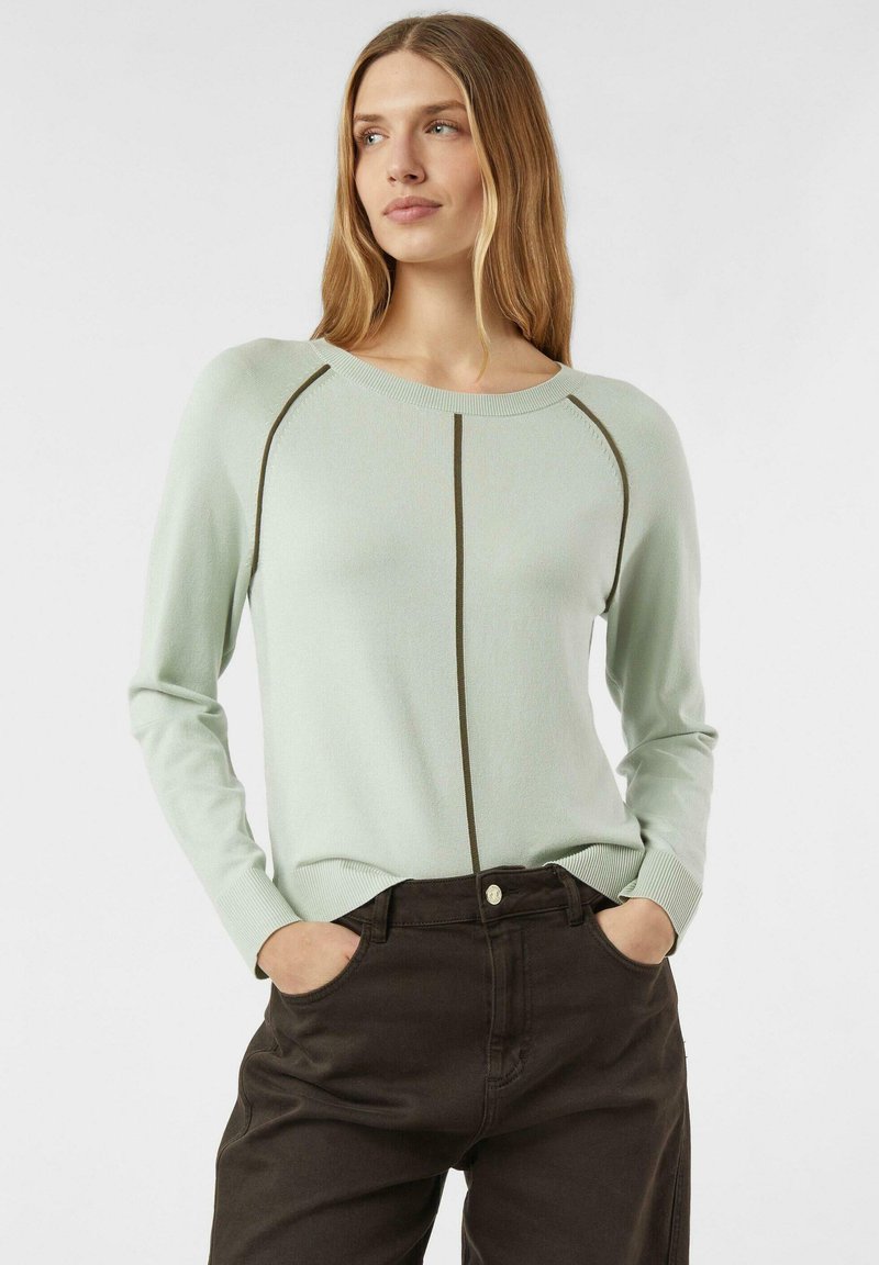 Kobieta o długich blond włosach, ubrana w jasnozielony sweter z ciemnymi pionowymi pasami i ciemnobrązowe spodnie, z rękami w kieszeniach, patrząca na bok.