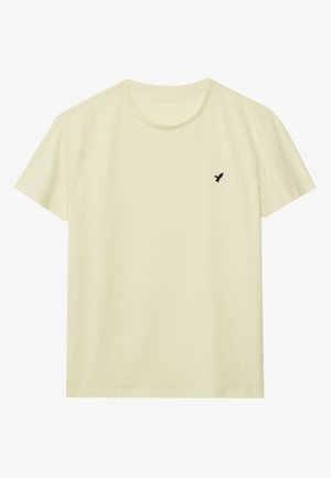 Camiseta de color amarillo claro con mangas cortas y cuello redondo, con un pequeño logo negro de pájaro en el pecho izquierdo, mostrada sobre un fondo blanco liso.