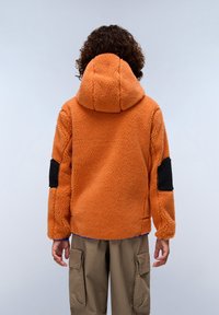 Oransje fleecejakke med hette, med svarte lappakenter på ermene. Teksturen er myk og fluffy; avslappet passform.