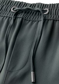 Shorts de sport vert foncé avec une taille élastique, un cordon de serrage ajustable et un tissu lisse et texturé. Présente des accents en métal.