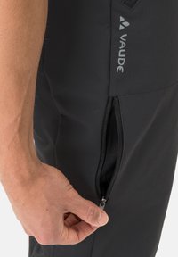Pantaloni neri da esterno con tasca con zip. Il materiale appare resistente e ha una texture. Il logo del marchio "Vaude" è visibile sul lato.